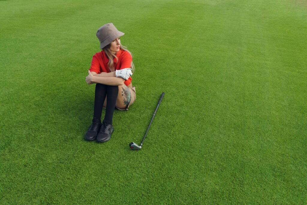 A woman in a red shirt sitting on green grass with a golf club nearby.